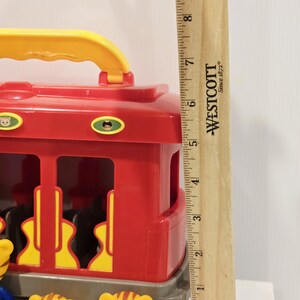 K&ouml;nnte beinhalten: Ein roter und gelber Spielzeugwagen mit gelbem Griff. Der Wagen hat gelbe Aussparungen und kleine Charakteraufkleber. Mehrere kleine gelbe Tigerfiguren befinden sich vor dem Wagen. Ein Holzlineal misst etwa 23 cm.