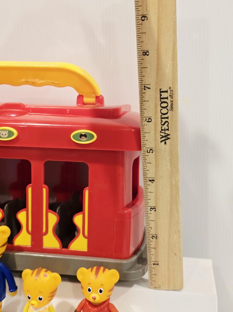 K&ouml;nnte beinhalten: Ein roter und gelber Spielzeugwagen mit gelbem Griff. Der Wagen hat gelbe Aussparungen und kleine Charakteraufkleber. Mehrere kleine gelbe Tigerfiguren befinden sich vor dem Wagen. Ein Holzlineal misst etwa 23 cm.