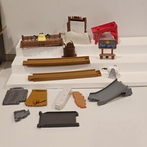 May include: A collection of toy car track pieces and accessories, including a yellow car, brown fences, a red sign that says "Fireball Beach", and a gray slide. The set is arranged on a white surface.