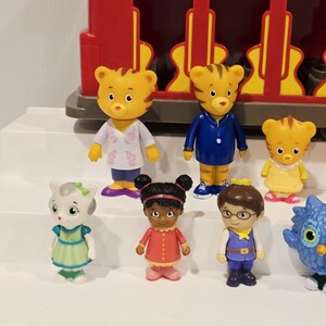K&ouml;nnte beinhalten: Eine Sammlung farbenfroher Daniel Tiger's Neighborhood-Figuren, darunter Daniel Tiger, Katerina Kittycat, O die Eule und andere. Die Figuren stehen vor einem roten Spielzeugzug. Die Charaktere tragen verschiedene Outfits.