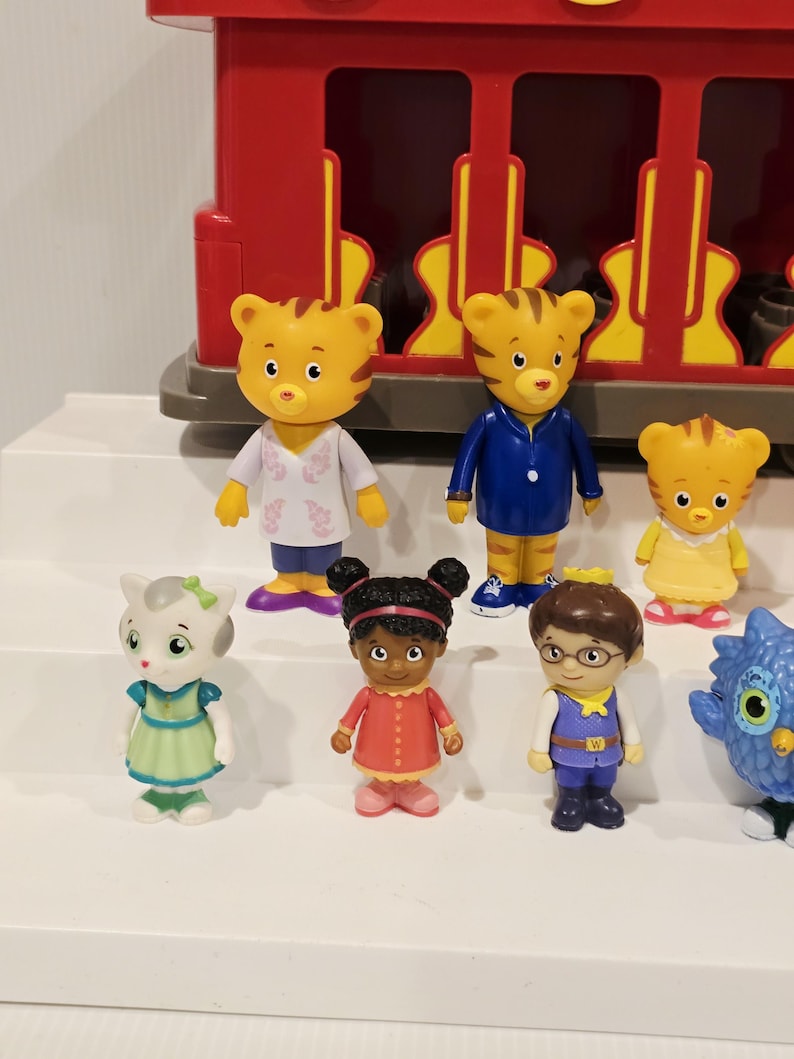K&ouml;nnte beinhalten: Eine Sammlung farbenfroher Daniel Tiger's Neighborhood-Figuren, darunter Daniel Tiger, Katerina Kittycat, O die Eule und andere. Die Figuren stehen vor einem roten Spielzeugzug. Die Charaktere tragen verschiedene Outfits.