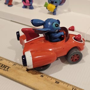 May include: A red toy car with white accents and a blue Stitch figurine in the driver's seat. Other Stitch figurines are visible in the background. The car has black wheels and red lights.