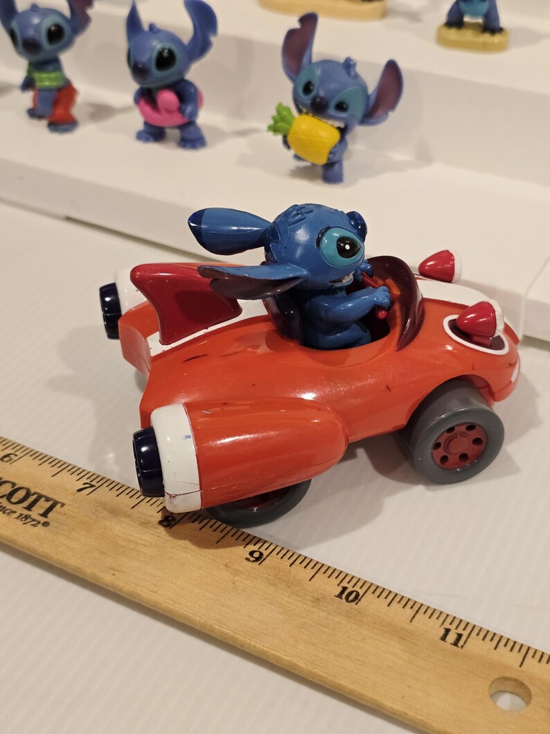 May include: A red toy car with white accents and a blue Stitch figurine in the driver's seat. Other Stitch figurines are visible in the background. The car has black wheels and red lights.