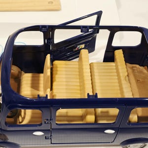 May include: A blue toy car with an open door, revealing tan seats and interior details. The car has a steering wheel and windows. The toy car is designed for children's play and imaginative scenarios.