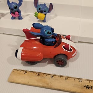 May include: A red toy car with white stripes and a blue character figure driving. The car has gray wheels and a fin. Other blue character figurines are on a white shelf in the background.