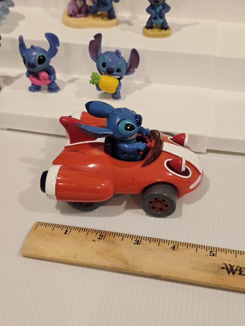 May include: A red toy car with white stripes and a blue character figure driving. The car has gray wheels and a fin. Other blue character figurines are on a white shelf in the background.
