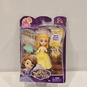 May include: A Sofia the First doll in a clear plastic package. The doll wears a yellow dress and has blonde hair. The packaging includes the text "#3 Bea a Good Sister" and the Sofia the First logo. A small green fan is also included.