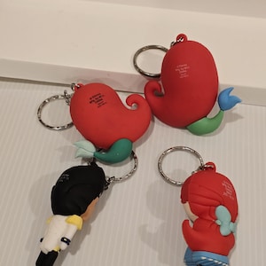 May include: Four keychains featuring characters from a Disney movie. The keychains have a red, blue, green, black, and white color scheme. The keychains are attached to silver keyrings. The keychains are made of a rubbery material.