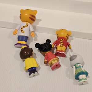 May include: A collection of small, colorful figurines featuring characters from a children's animated series. The figures include a tiger, a girl with black hair, and other characters in various outfits. The figurines are made of plastic and are brightly colored.