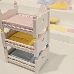 Puede incluir: Una cama litera blanca en miniatura con tres niveles, cada uno con una almohada de diferente color: rosa, amarillo y azul. La cama tiene recortes decorativos en forma de coraz&oacute;n y est&aacute; acentuada con detalles rosas, amarillos y azules.