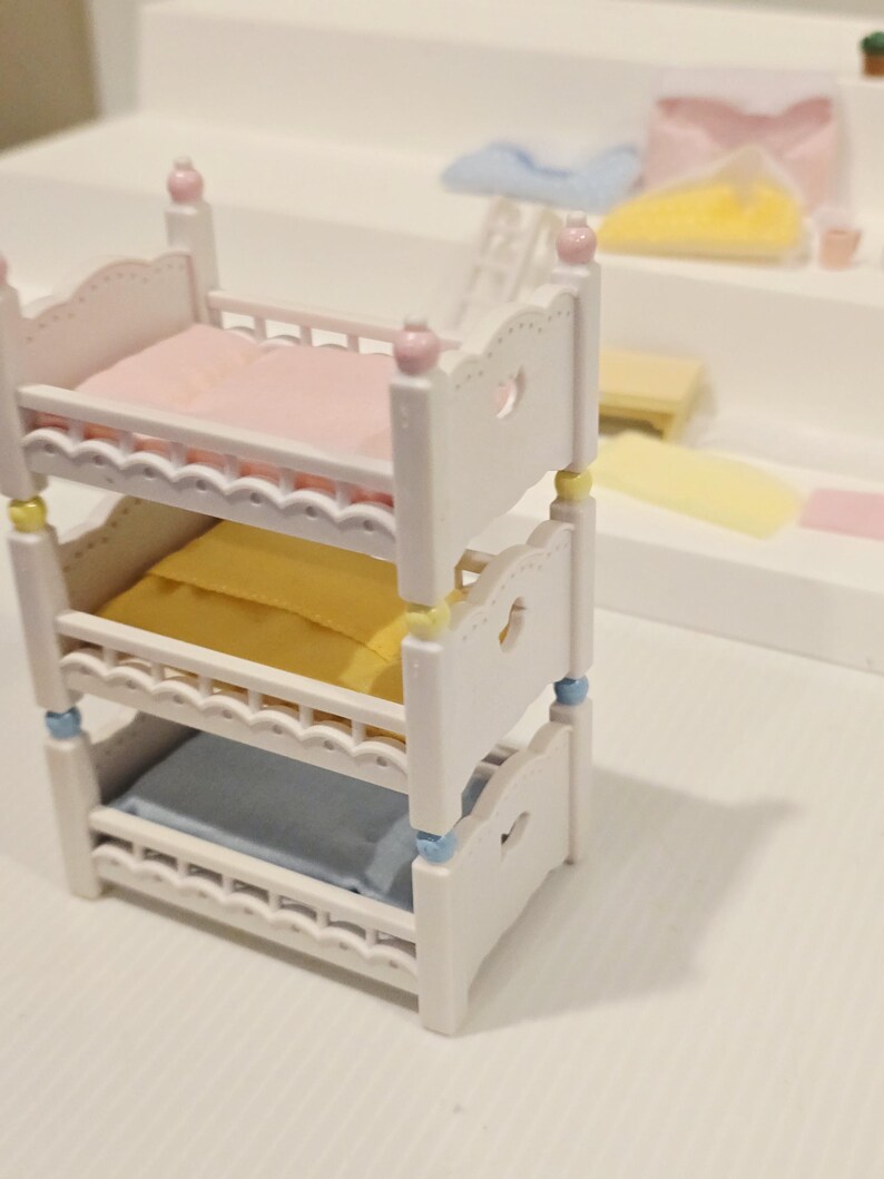 Puede incluir: Una cama litera blanca en miniatura con tres niveles, cada uno con una almohada de diferente color: rosa, amarillo y azul. La cama tiene recortes decorativos en forma de coraz&oacute;n y est&aacute; acentuada con detalles rosas, amarillos y azules.
