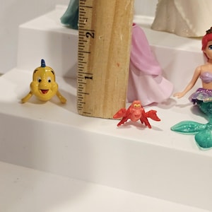 K&ouml;nnte beinhalten: Eine gelbe Cartoon-Fischfigur mit blauer Flosse und eine rote Krabbenfigur werden auf einer wei&szlig;en Oberfl&auml;che gezeigt. Ein Holzlineal wird neben den Figuren platziert. Weitere Prinzessinnenfiguren sind im Hintergrund zu sehen.