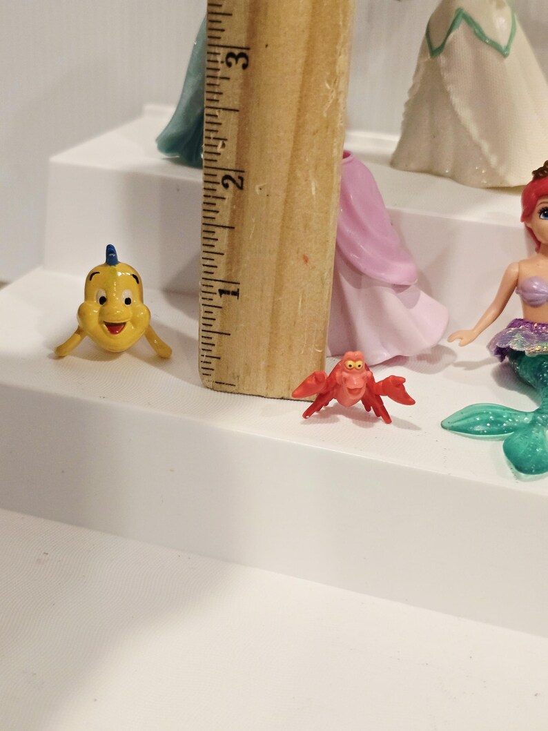 K&ouml;nnte beinhalten: Eine gelbe Cartoon-Fischfigur mit blauer Flosse und eine rote Krabbenfigur werden auf einer wei&szlig;en Oberfl&auml;che gezeigt. Ein Holzlineal wird neben den Figuren platziert. Weitere Prinzessinnenfiguren sind im Hintergrund zu sehen.