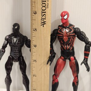 May include: Two Spider-Man action figures, one black and one red and black, stand next to a wooden ruler. The black figure has a white spider emblem. The red and black figure has a spider web design. The ruler measures up to 7 inches.