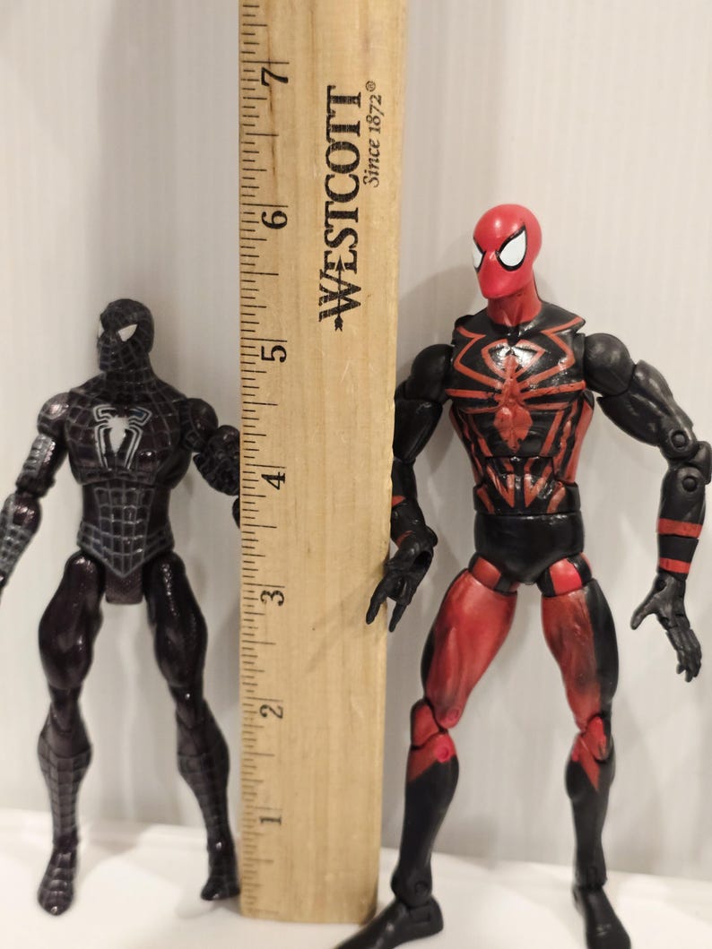May include: Two Spider-Man action figures, one black and one red and black, stand next to a wooden ruler. The black figure has a white spider emblem. The red and black figure has a spider web design. The ruler measures up to 7 inches.
