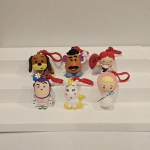 May include: Seven character keyrings with red loops. The keyrings depict cartoon characters such as a dog, Mr. Potato Head, a cowgirl, a princess, a unicorn, and a blonde girl in a pink hood. They are arranged on a white stepped surface.