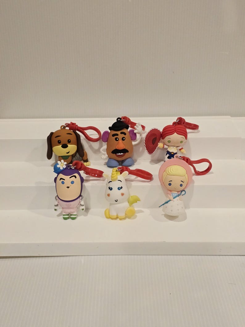 May include: Seven character keyrings with red loops. The keyrings depict cartoon characters such as a dog, Mr. Potato Head, a cowgirl, a princess, a unicorn, and a blonde girl in a pink hood. They are arranged on a white stepped surface.