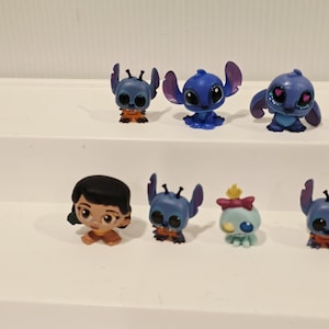 May include: A collection of small, colorful figurines featuring characters from the animated film "Lilo & Stitch." The figurines include Stitch in various poses, along with Lilo and another character. The figures are primarily blue, orange, and tan.