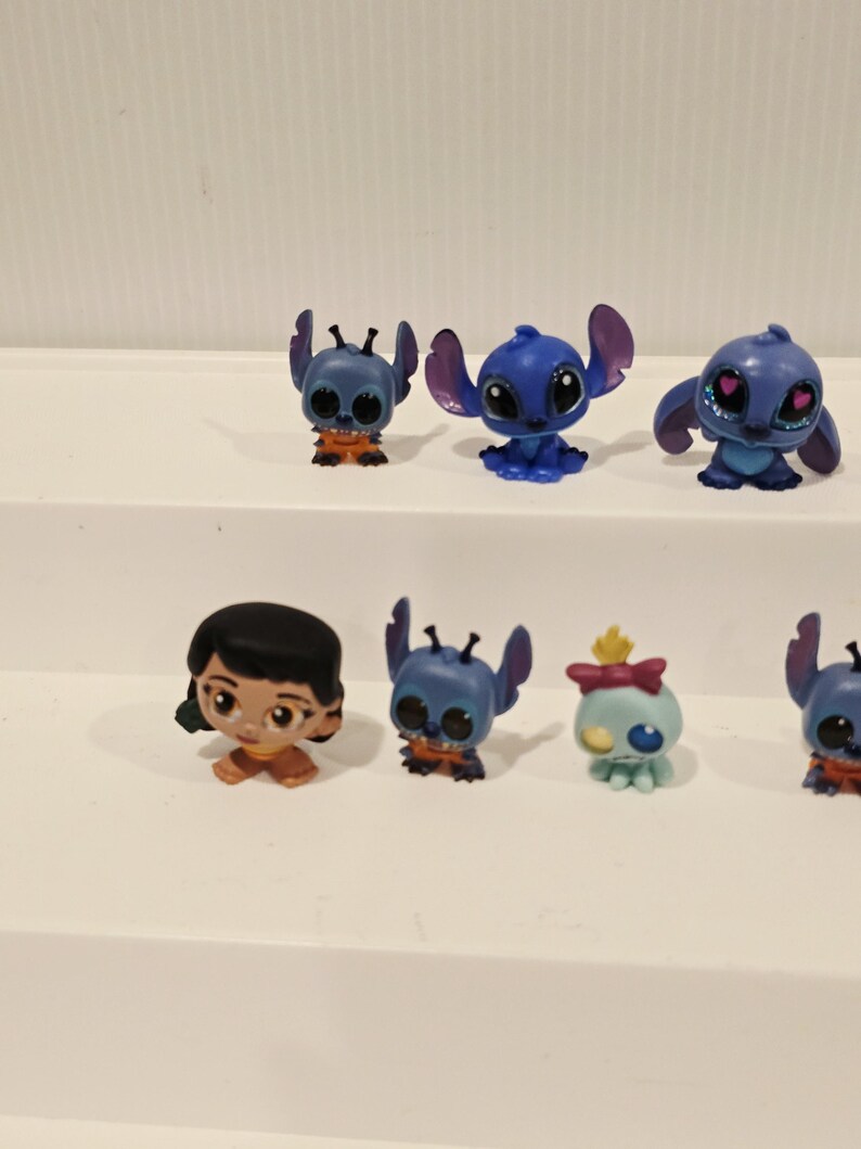May include: A collection of small, colorful figurines featuring characters from the animated film "Lilo & Stitch." The figurines include Stitch in various poses, along with Lilo and another character. The figures are primarily blue, orange, and tan.