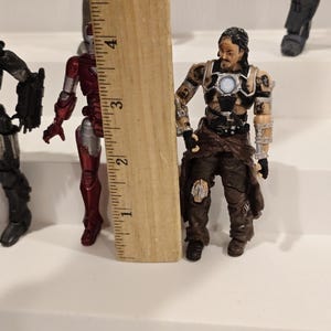 May include: A collection of action figures, including a red and silver Iron Man figure and a figure with brown and black clothing. A wooden ruler is placed next to the figures, measuring approximately 3 inches.