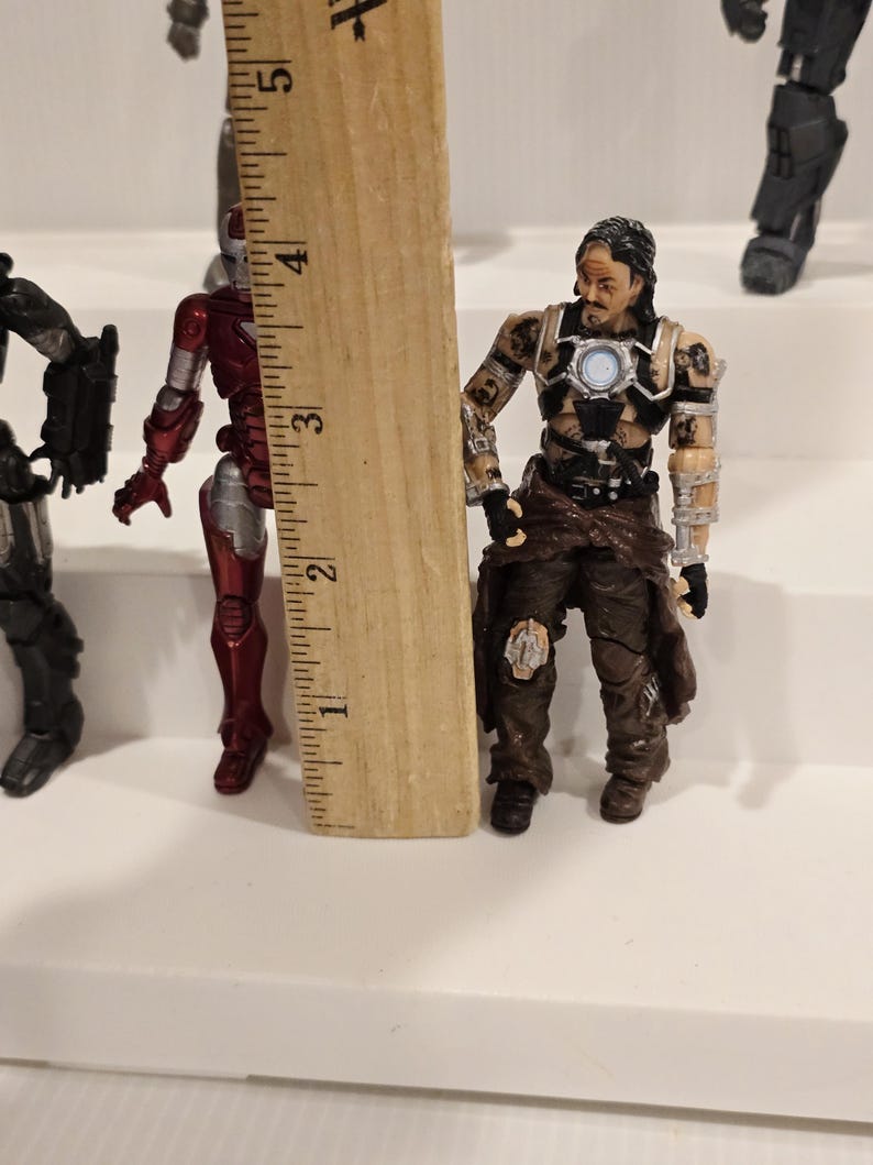 May include: A collection of action figures, including a red and silver Iron Man figure and a figure with brown and black clothing. A wooden ruler is placed next to the figures, measuring approximately 3 inches.