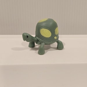 May include: A small, green turtle figurine with yellow accents on its shell and head. The toy turtle has four stubby legs and a small head, and is set against a plain white background.