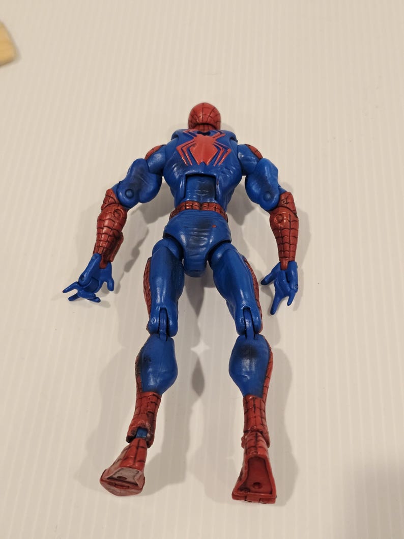 Pode incluir: Figura de a&ccedil;&atilde;o azul e vermelha de um super-her&oacute;i, vista por tr&aacute;s. A figura tem um emblema de aranha vermelho nas costas e detalhes vermelhos nos bra&ccedil;os e pernas. A figura est&aacute; em pose com os bra&ccedil;os ao lado.
