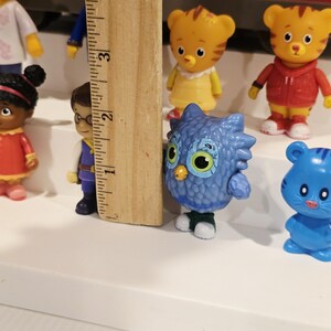 K&ouml;nnte beinhalten: Eine Sammlung farbenfroher Daniel Tiger's Neighborhood-Figuren, darunter Daniel Tiger, Katerina Kittycat, O die Eule und andere. Die Figuren sind aus Kunststoff und etwa 5 cm gro&szlig;. Ein Holzlineal befindet sich im Bild.