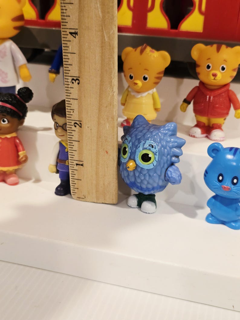 K&ouml;nnte beinhalten: Eine Sammlung farbenfroher Daniel Tiger's Neighborhood-Figuren, darunter Daniel Tiger, Katerina Kittycat, O die Eule und andere. Die Figuren sind aus Kunststoff und etwa 5 cm gro&szlig;. Ein Holzlineal befindet sich im Bild.