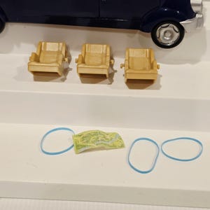 May include: A dark blue toy car with three small tan armchairs, a miniature map, and three blue rubber bands. The car has a rounded shape and visible wheels. The armchairs are arranged in a row.