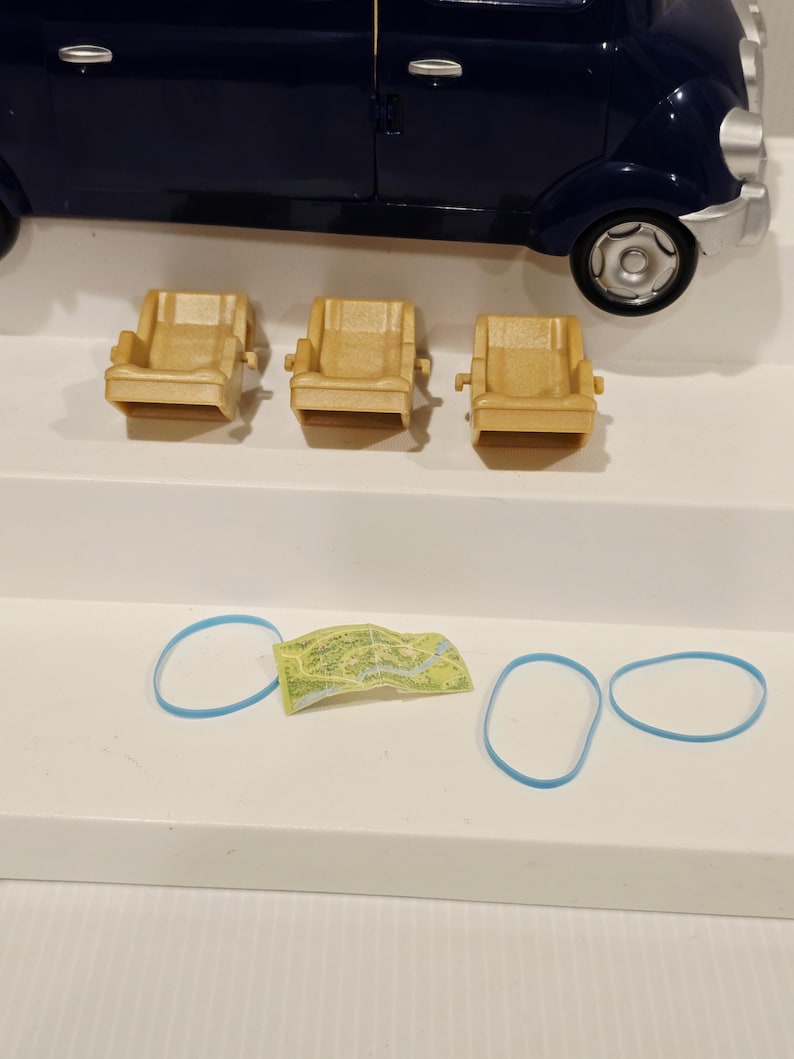 May include: A dark blue toy car with three small tan armchairs, a miniature map, and three blue rubber bands. The car has a rounded shape and visible wheels. The armchairs are arranged in a row.