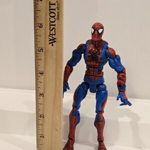 Pode incluir: Figura de a&ccedil;&atilde;o do Homem-Aranha com seu traje vermelho e azul cl&aacute;ssico, com um emblema de aranha preta no peito. A figura est&aacute; ao lado de uma r&eacute;gua de madeira, marcada com o logotipo "WESTCOTT Since 1872".