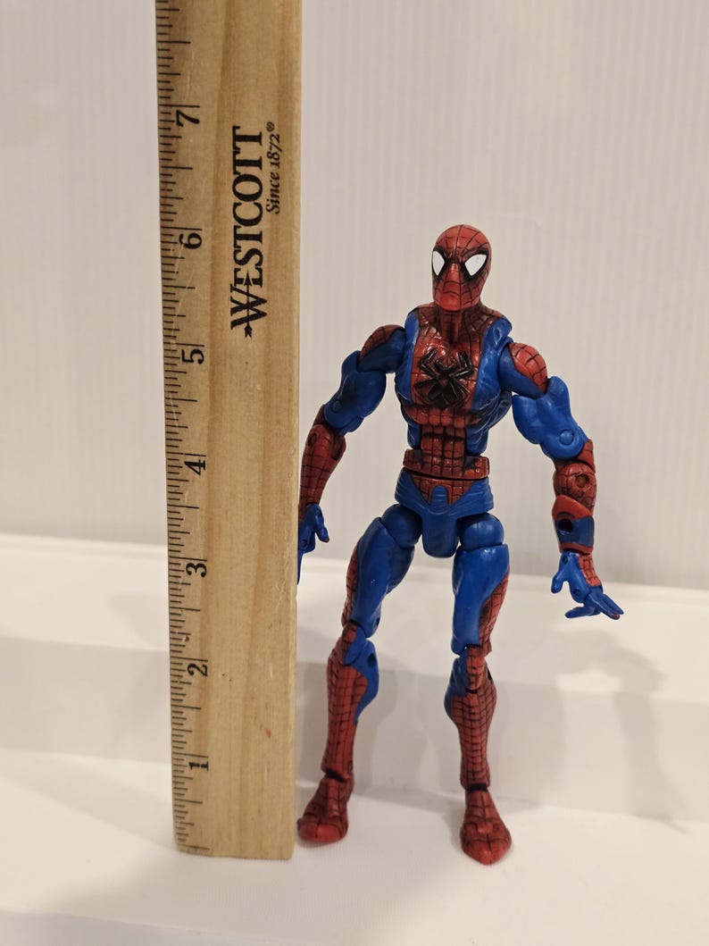 Pode incluir: Figura de a&ccedil;&atilde;o do Homem-Aranha com seu traje vermelho e azul cl&aacute;ssico, com um emblema de aranha preta no peito. A figura est&aacute; ao lado de uma r&eacute;gua de madeira, marcada com o logotipo "WESTCOTT Since 1872".