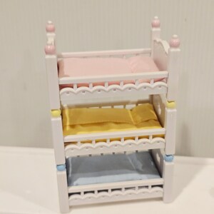 Puede incluir: Una cama triple en miniatura de color blanco con ropa de cama rosa, amarilla y azul. La cama tiene detalles decorativos y remates redondeados. La ropa de cama incluye almohadas y mantas.