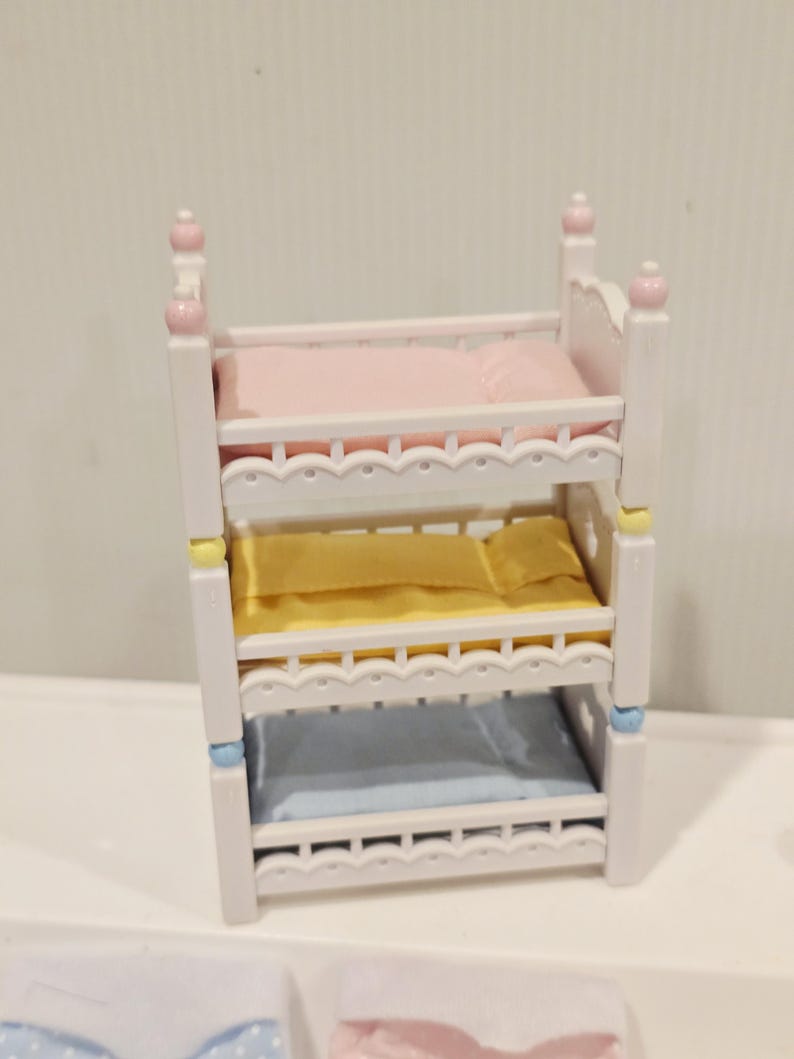 Puede incluir: Una cama triple en miniatura de color blanco con ropa de cama rosa, amarilla y azul. La cama tiene detalles decorativos y remates redondeados. La ropa de cama incluye almohadas y mantas.