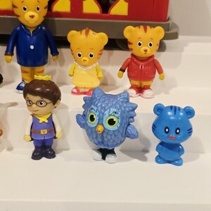 K&ouml;nnte beinhalten: Eine Sammlung von Daniel Tiger's Neighborhood-Figuren. Die Figuren zeigen Daniel Tiger, Eule, Katerina Kittycat und andere Charaktere. Die Spielzeuge sind farbenfroh und aus Kunststoff. Ein roter Zugwagen ist im Hintergrund zu sehen.