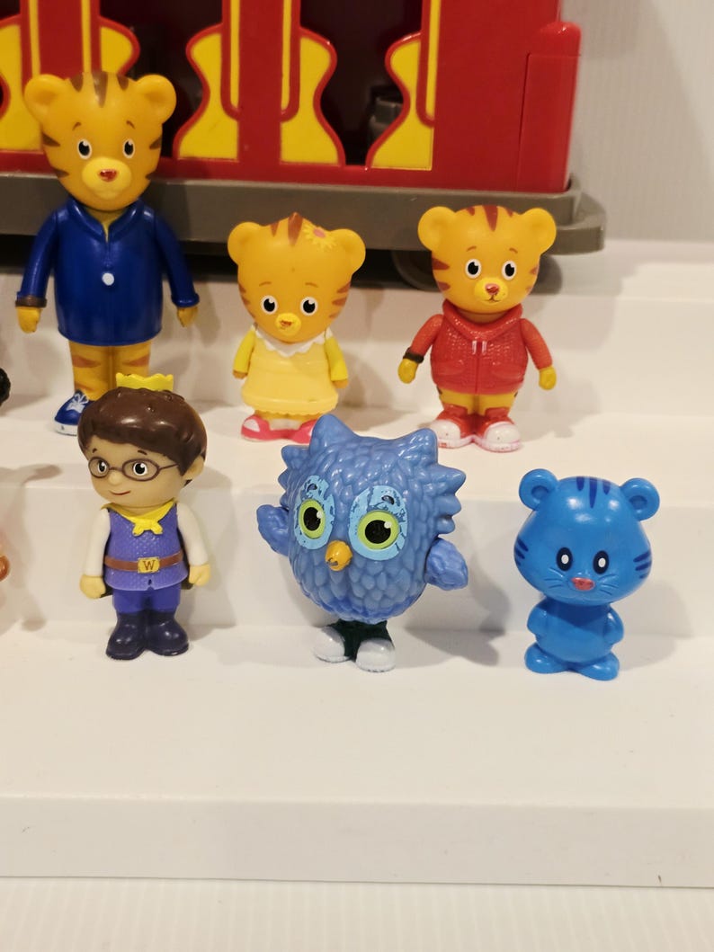 K&ouml;nnte beinhalten: Eine Sammlung von Daniel Tiger's Neighborhood-Figuren. Die Figuren zeigen Daniel Tiger, Eule, Katerina Kittycat und andere Charaktere. Die Spielzeuge sind farbenfroh und aus Kunststoff. Ein roter Zugwagen ist im Hintergrund zu sehen.