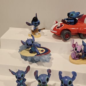 May include: A collection of Stitch figurines in various poses. One figure plays a guitar, another surfs, and another drives a red car. Other figures are standing. The figurines are blue, with some wearing outfits.