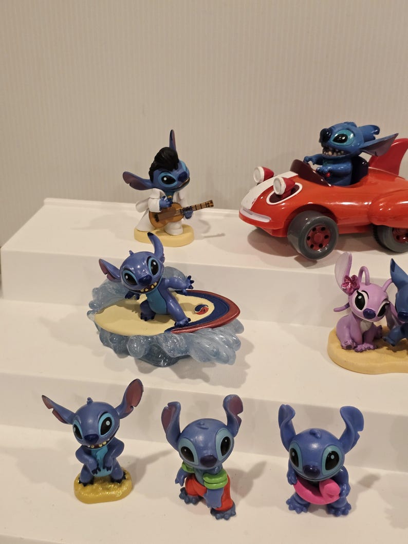 May include: A collection of Stitch figurines in various poses. One figure plays a guitar, another surfs, and another drives a red car. Other figures are standing. The figurines are blue, with some wearing outfits.