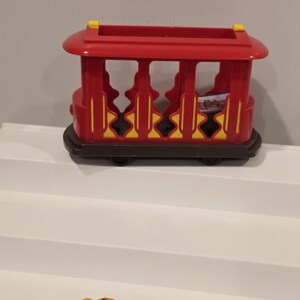 May include: A red and yellow toy train car with a brown base. The toy train car has a rectangular shape with open sides and a rounded roof. The wheels are black. The toy is on a white surface.