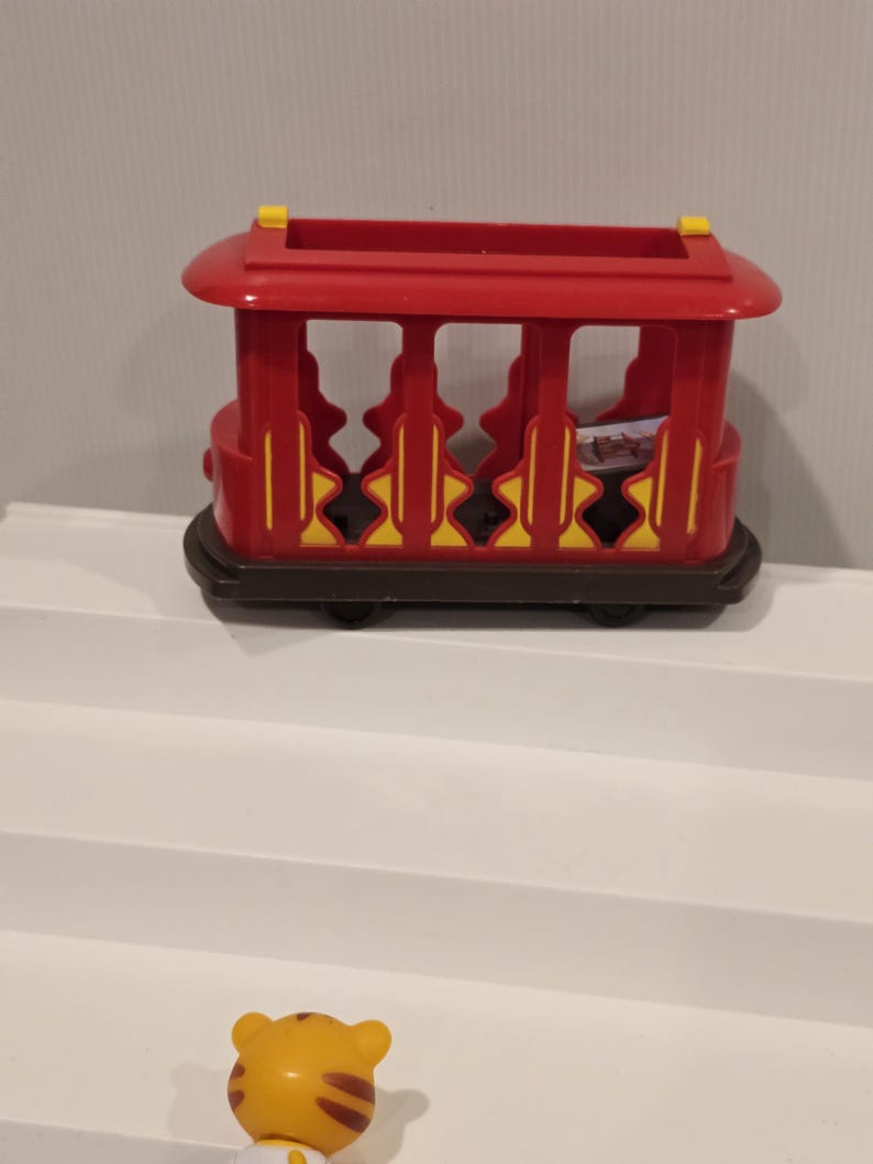May include: A red and yellow toy train car with a brown base. The toy train car has a rectangular shape with open sides and a rounded roof. The wheels are black. The toy is on a white surface.