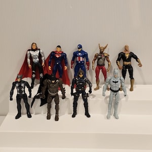May include: A collection of superhero action figures, including Superman, Captain America, and Batman, are arranged on a white stepped surface. The figures are brightly colored and detailed, showcasing various costumes and poses. The figures are approximately 6 inches tall.