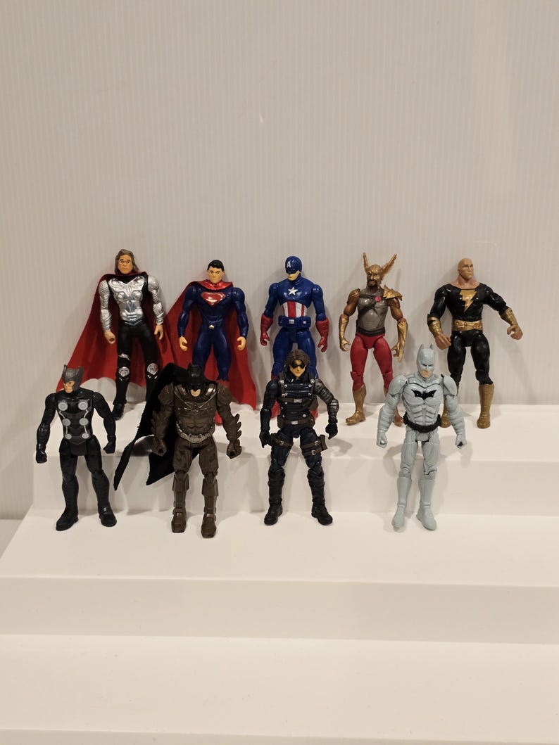 May include: A collection of superhero action figures, including Superman, Captain America, and Batman, are arranged on a white stepped surface. The figures are brightly colored and detailed, showcasing various costumes and poses. The figures are approximately 6 inches tall.