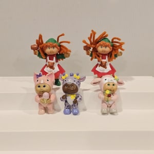 May include: A collection of six vintage Cabbage Patch Kids figurines. Two figures are dressed as elves, and the other four are in animal costumes: a pig, a giraffe, and two bunnies. The figurines are in various colors, including red, green, pink, and brown.