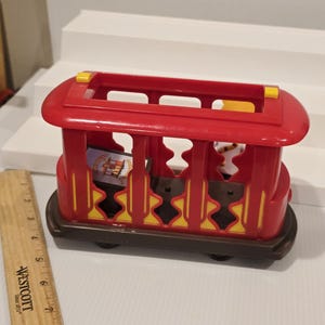 May include: A red and brown toy trolley with yellow accents. The trolley has a rectangular shape with open sides and a roof. The wheels are black. The trolley appears to be made of plastic and is designed for children's play.
