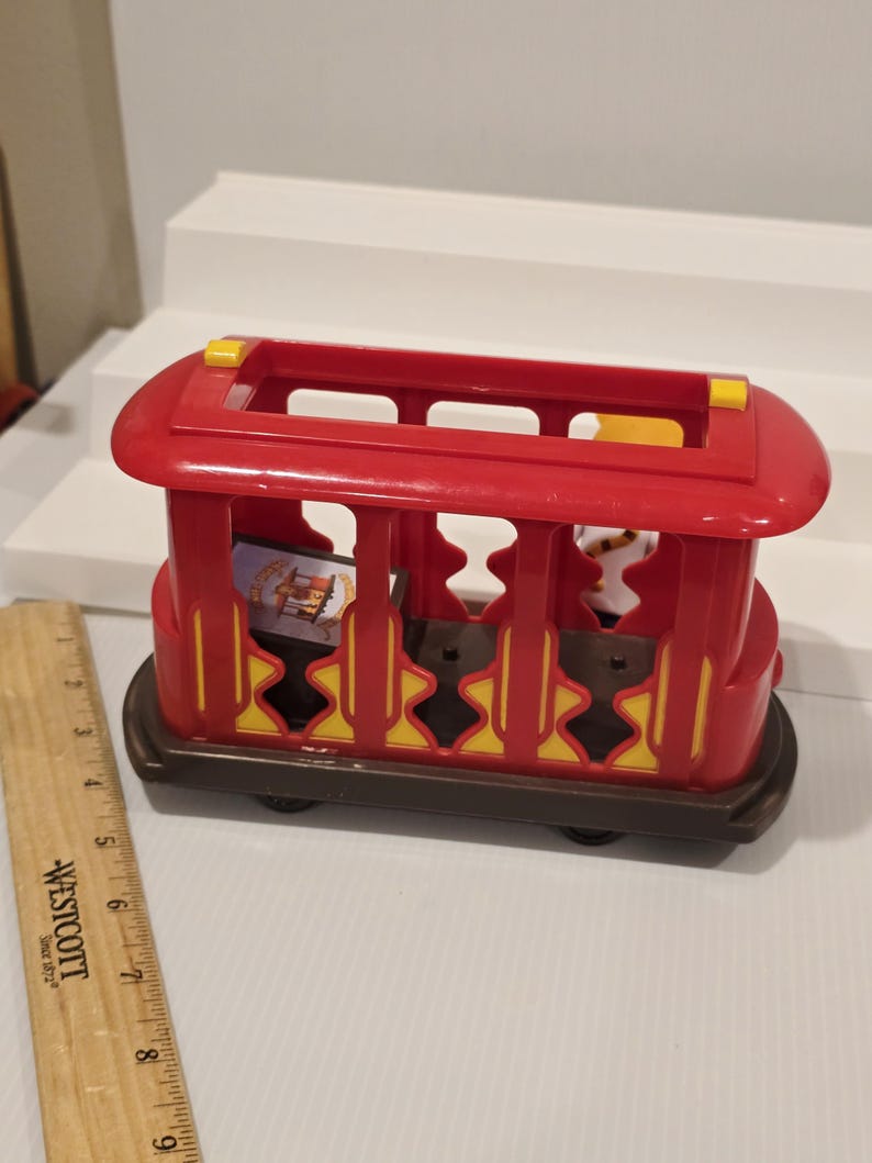 May include: A red and brown toy trolley with yellow accents. The trolley has a rectangular shape with open sides and a roof. The wheels are black. The trolley appears to be made of plastic and is designed for children's play.