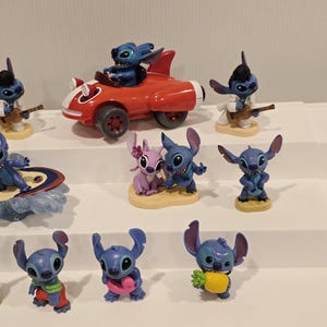 May include: A collection of blue alien character figurines in various poses. One figurine is in a red race car, another is playing a guitar, and others are holding items like a pineapple and a pink heart. The figurines are displayed on white shelves.