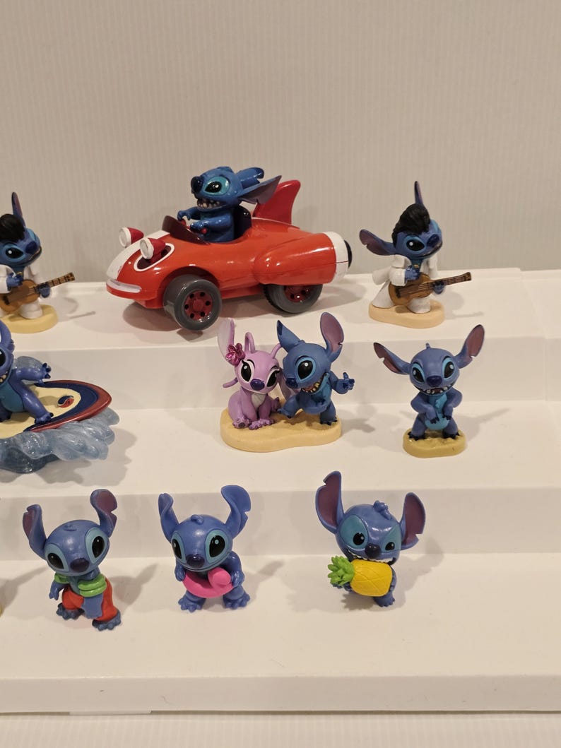 May include: A collection of blue alien character figurines in various poses. One figurine is in a red race car, another is playing a guitar, and others are holding items like a pineapple and a pink heart. The figurines are displayed on white shelves.
