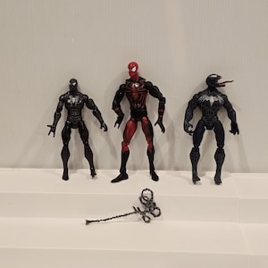 May include: Three Spider-Man action figures in black, red, and blue. The figures are standing on a white surface. A silver-colored weapon lies in front of the figures. The figures are detailed with various textures and designs.