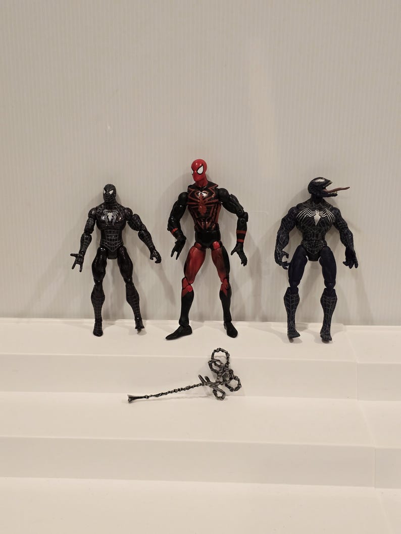 May include: Three Spider-Man action figures in black, red, and blue. The figures are standing on a white surface. A silver-colored weapon lies in front of the figures. The figures are detailed with various textures and designs.